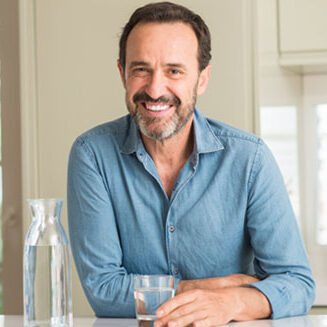 Middle age man drinking a glass of water with a happy face standing and smiling with a confident smile showing teeth