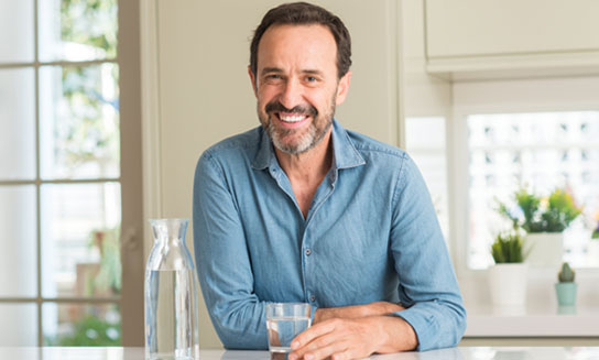 Middle age man drinking a glass of water with a happy face standing and smiling with a confident smile showing teeth